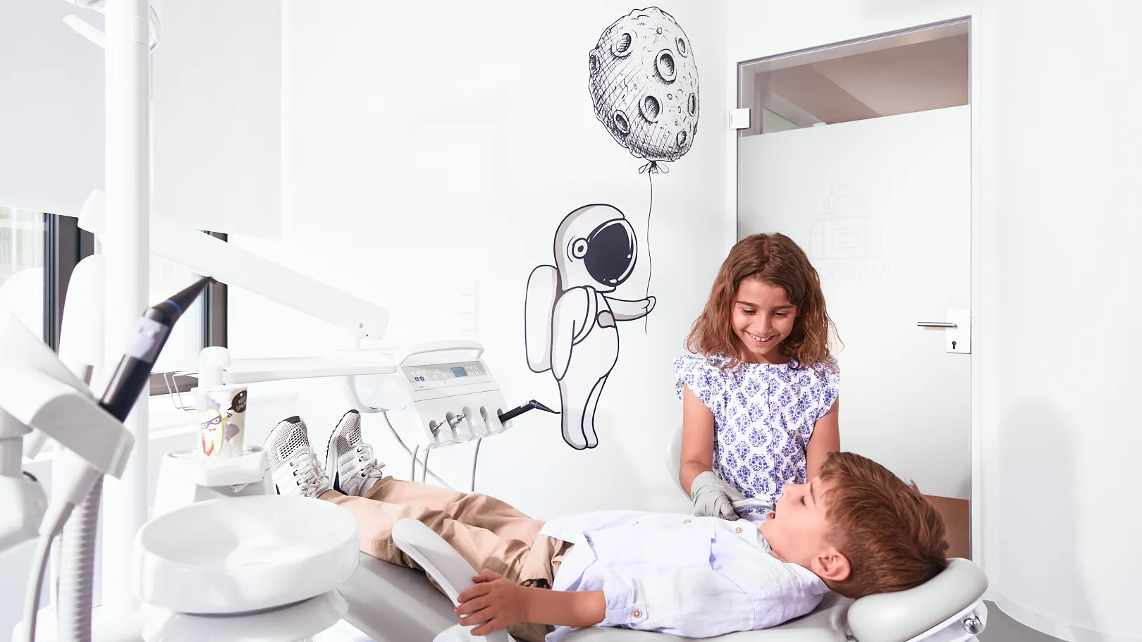 Kinderzahnheilkunde von Anfang an – RKM 740 Zahnmedizin Düsseldorf Zahnärztliche Behandlungssituation im RKM 740 Düsseldorf: zwei Kinder im hellen Praxisraum mit Mond-Ballon-Motiv – Symbol für Vertrauen, Neugier und behutsame Kinderzahnheilkunde von Anfang an